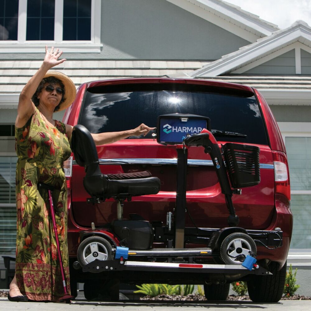 A woman in a floral dress waves beside a red van equipped with the Harmar Heavy Duty Fusion Lift (AL300HD), which securely carries a mobility scooter mounted at the rear of the vehicle.