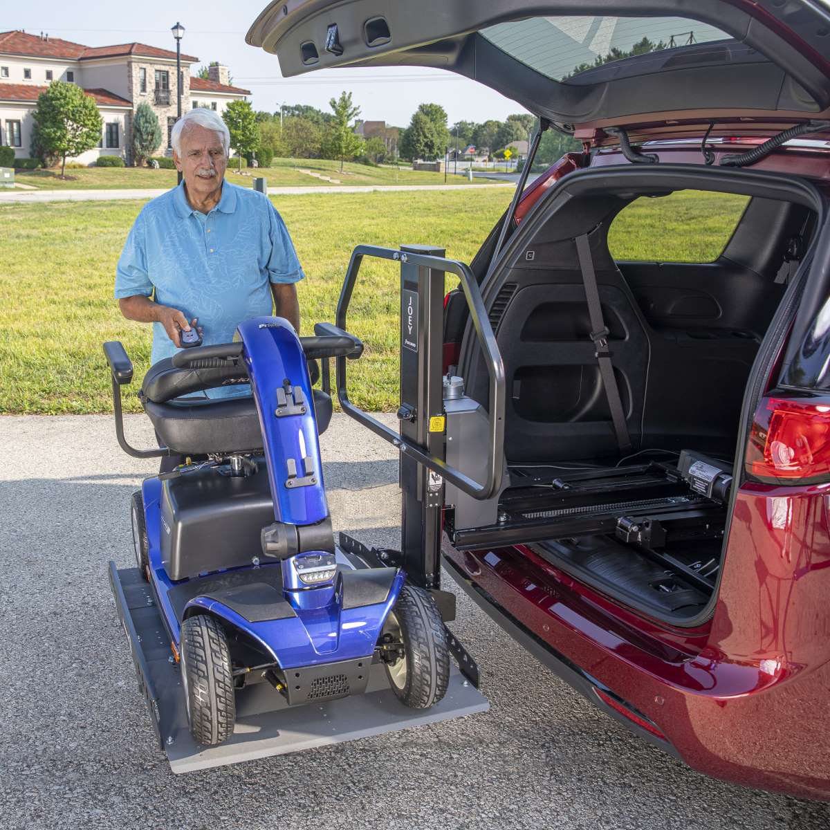 An older man stands beside a blue mobility scooter as a Bruno Joey Platform Style Interior Lift (VSL-4400) lifts it on a platform into the back of a red vehicle.