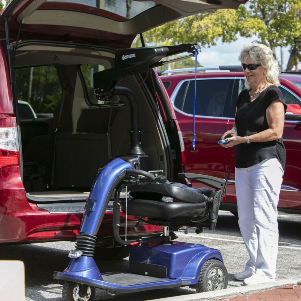 A woman uses a remote control to operate a Harmar Premium Inside Lift (AL435), loading a blue mobility scooter into the back of a red minivan in a parking lot.
